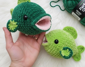 May include: Two handmade crochet fish in shades of green. One is a darker green with a pink mouth, and the other is a lighter green. Both have black eyes and are made with a soft, textured yarn. The fish are displayed with green yarn.