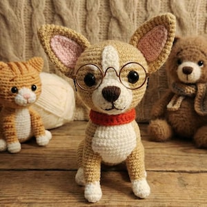 Op de afbeelding: Een gehaakte hond met een bril en een rode halsband is het middelpunt van deze afbeelding. Een gehaakte kat en teddybeer zijn ook zichtbaar. De hond is bruin en wit, met grote oren en een bruine neus.