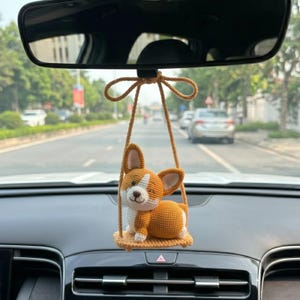 Puede incluir: Un adorno de coche de perro corgi tejido a ganchillo en tonos naranja, blanco y marrón, colgado del espejo retrovisor de un coche. El perro está sentado en un pequeño columpio, añadiendo un toque lúdico al interior del coche.