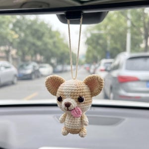 May include: A small, crocheted dog ornament in shades of cream and light brown, featuring large dark eyes, a brown nose, and a small pink tongue sticking out. The figure is suspended by a thin, light-coloured string, designed for hanging display.