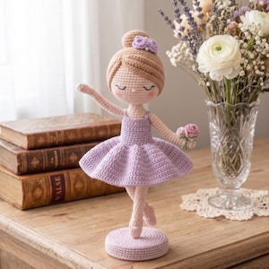May include: A crocheted ballerina doll with light brown hair in a bun, wearing a lavender dress and matching ballet slippers. The doll has closed eyes, pink cheeks, and holds a small pink flower. It stands on a circular base.
