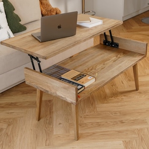 Puede incluir: Mesa de centro elevable de madera clara con ordenador portátil y libros. La mesa está en un salón con un sofá beige, cojines decorativos y un oso de peluche. La mesa mide 102 cm de largo.