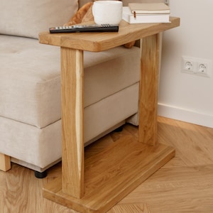 May include: A natural wood side table with a rectangular top and two vertical supports. A remote, books, and a white mug sit on the table. A beige sofa, a teddy bear, and a decorative tree pillow are in the background.