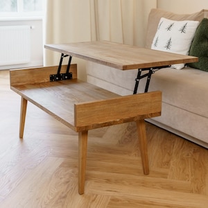 May include: A wooden lift-top coffee table with a light brown finish. The table is in the raised position, revealing the black metal lifting mechanism. The table is in a living room setting with a beige sofa and hardwood flooring.