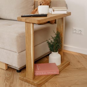 Puede incluir: Una mesa auxiliar de madera con una parte superior rectangular y dos soportes verticales. Una taza de cerámica blanca, libros y un mando a distancia están sobre la mesa. Un pequeño jarrón con vegetación y un libro titulado "NATURAL SPACES".