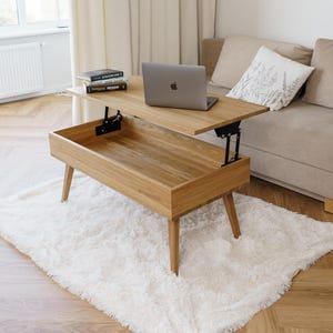 Könnte beinhalten: Ein helles Holz-Couchtisch mit Hubtischplatte, Laptop und Büchern. Der Tisch steht auf einem weißen, flauschigen Teppich, mit einem beigen Sofa und Fenster im Hintergrund. Die Tischplatte ist angehoben und gibt Stauraum frei.