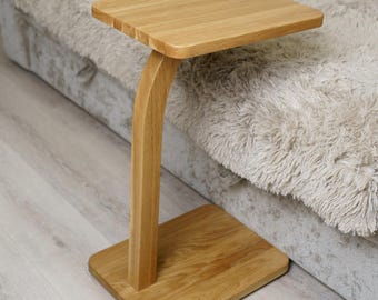 May include: A wooden side table with a square top and a curved leg. The table is made of light-colored wood and has a rectangular base. The table is positioned next to a gray sofa with a fluffy blanket and an orange pillow.