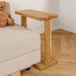 May include: A wooden side table with a rectangular top and a single leg. The table is made of light-colored wood and is positioned next to a beige sofa. A brown teddy bear sits on the sofa. The table's dimensions are approximately 24 inches long, 12 inches wide, and 26 inches high.