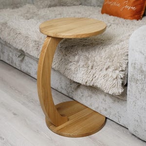 Op de afbeelding: Een houten bijzettafel met een rond blad en een gebogen steun. De tafel is gemaakt van lichtgekleurd hout en heeft een cirkelvormige basis. De tafel staat naast een grijze bank met een zachte deken en een oranje kussen. De tafel is ongeveer 60 cm hoog.