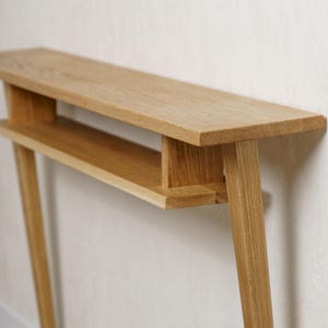 Può includere: Una consolle in legno di rovere chiaro dal design minimalista. Il tavolo presenta un piano lungo e rettangolare con un ripiano inferiore e gambe angolate. La venatura naturale del legno è visibile e il tavolo è appoggiato a un muro neutro.