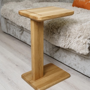May include: A wooden side table with a square top and base, connected by a rectangular support. The table is made of light-colored wood and stands on a light-colored wooden floor. The table's height is approximately 24 inches.