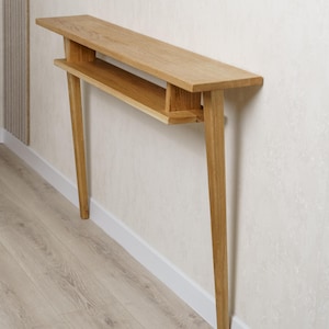 Puede incluir: Una consola de madera clara con un diseño minimalista. La mesa tiene una parte superior larga y rectangular con un estante inferior y patas anguladas. La mesa mide aproximadamente 122 cm de largo.