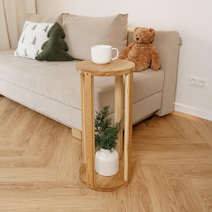 Puede incluir: Una pequeña mesa auxiliar redonda de madera con una taza blanca encima. Un pequeño jarrón blanco con vegetación se encuentra dentro de la mesa. Un sofá beige con almohadas y un oso de peluche están en el fondo. La habitación tiene paredes claras y suelo de madera.