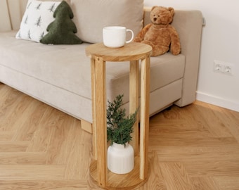 Könnte beinhalten: Kleiner, runder Beistelltisch aus Holz mit einer weißen Tasse und einer kleinen Pflanze in einer weißen Vase. Im Hintergrund ein beiges Sofa mit Zierkissen und einem Teddybär. Der Tisch ist etwa 61 cm hoch.