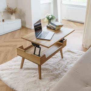 Könnte beinhalten: Ein hellbrauner, hölzerner Couchtisch mit Hubtischplatte und einem Laptop. Der Tisch steht auf einem weißen, flauschigen Teppich. Bücher sind auf dem Tisch gestapelt. Der Raum ist gut beleuchtet.