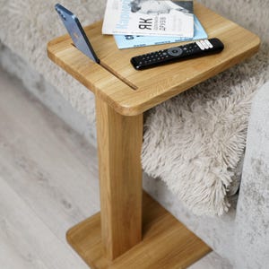 Könnte beinhalten: Ein helles Holz-Beistelltisch mit quadratischer Oberseite und einem einzelnen Bein. Ein Smartphone befindet sich in einem Schlitz auf dem Tisch, mit einem Buch und einer Fernbedienung. Der Tisch steht neben einem Sofa.