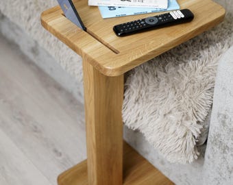 May include: A light-colored wooden side table with a square top and a single leg. A smartphone is in a slot on the table, with a book and remote control on top. The table is next to a couch.