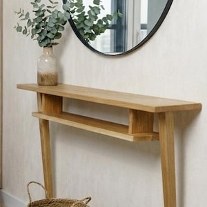 Puede incluir: Un salón moderno con una consola de madera, un espejo redondo negro y dos cestas tejidas. También se ve una lámpara de pie con una pantalla blanca y un sofá gris con cojines y una manta.