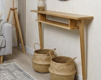 May include: A modern living room scene featuring a light wood console table with a shelf, a round black-framed mirror, and two woven baskets. A wooden tripod floor lamp with a white shade stands near a gray sofa with pillows and a throw blanket. A vase of greenery sits on the console.