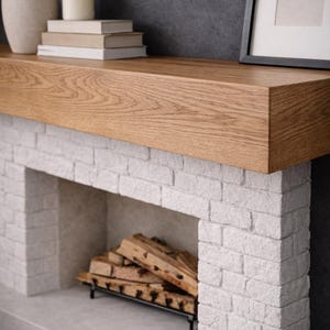 May include: A contemporary fireplace with a light oak mantel and a white brick facade. The hearth has a black metal grate filled with firewood. A vase, candle, and books sit on the mantel, with a framed picture on the right.