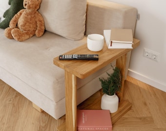 May include: A wooden side table with a built-in shelf, holding a white ceramic cup, books, and a small plant in a white vase. A remote control rests on the table's surface. A plush teddy bear and decorative pillows are on the adjacent beige sofa.