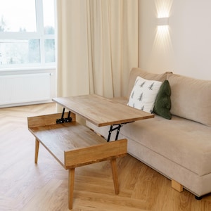 May include: A wooden lift-top coffee table with a lower shelf, set in a living room. The table is light brown and has a rectangular top. A beige sofa with decorative pillows is to the right. The room has light-colored walls, curtains, and a herringbone wood floor.