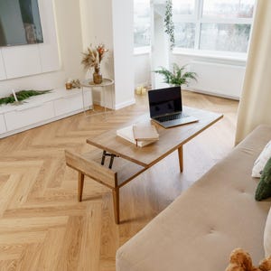 May include: A light brown wooden coffee table with a lift-top design, a laptop, and books. The table is in a living room with a beige sofa, hardwood floors, and a window.