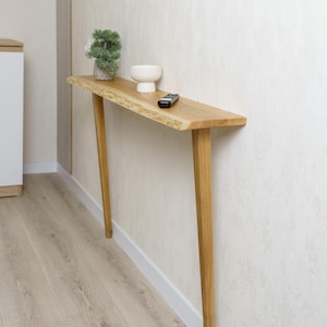 May include: A natural wood console table with a live edge top and angled legs. A small white bowl, a remote control, and a small potted plant sit on the table. The table is against a textured white wall.
