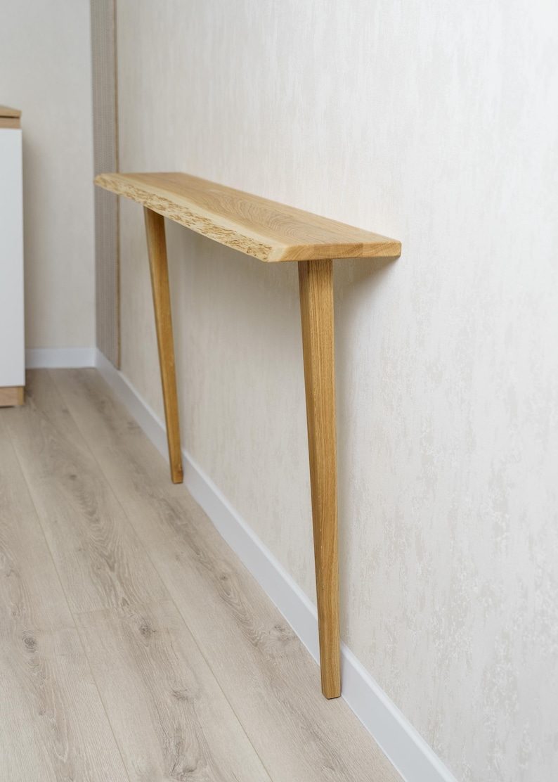 May include: A light-colored wooden console table with a live edge top and angled legs. The table is made of natural wood and is mounted against a wall. The legs are tapered and the table is approximately 48 inches long.