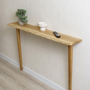May include: A narrow, light-colored wooden console table with a live edge. The table has four slender legs and is topped with a small plant, a white bowl, and a remote control. The table is approximately 48 inches long.