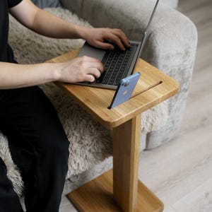 May include: A light-coloured wooden laptop table with a slot for a phone. The table has a rectangular top and a single leg with a square base. A laptop and a blue phone are on the table.