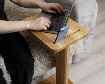 May include: A light-colored wooden laptop table with a slot for a phone. The table has a rectangular top and a single leg with a square base. A laptop and a blue phone are on the table.
