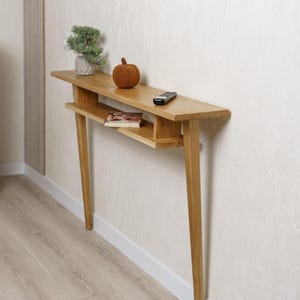 Op de afbeelding: Een houten console met een natuurlijke afwerking. Het heeft een lange, smalle bovenkant met een onderste plank en vier schuine poten. Een kleine plant, een decoratieve pompoen, een afstandsbediening en een boek liggen op de tafel.