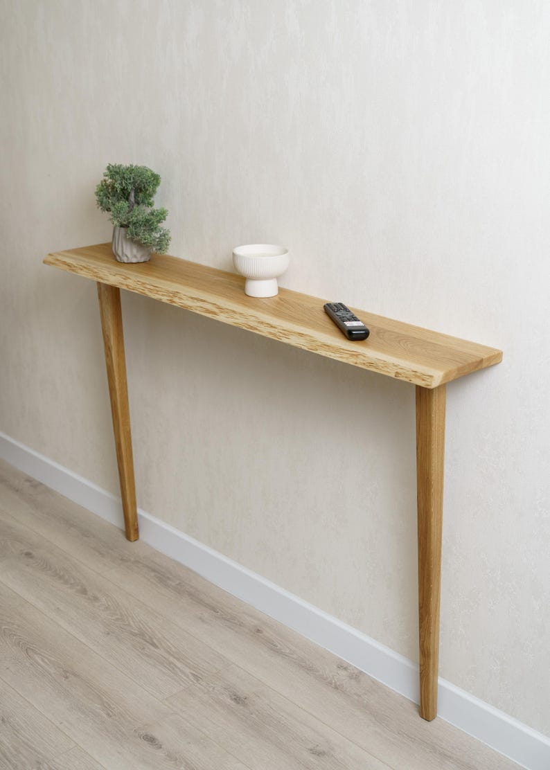 May include: A narrow, light-colored wooden console table with a live edge. The table has four slender legs and is topped with a small plant, a white bowl, and a remote control. The table is approximately 48 inches long.