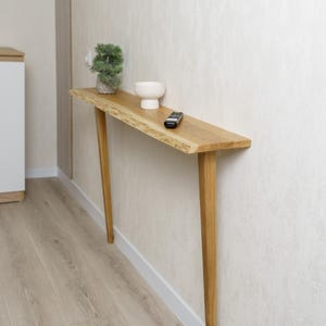 May include: A light-colored wooden console table with a live edge top and angled legs. A small white bowl, a remote control, and a small potted plant sit on the table. The table is against a textured wall.