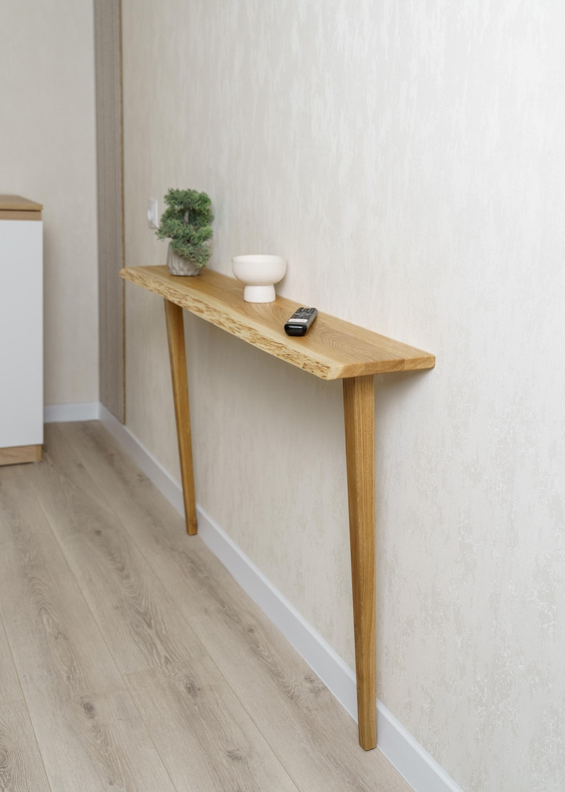 May include: A light-colored wooden console table with a live edge top and angled legs. A small white bowl, a remote control, and a small potted plant sit on the table. The table is against a textured wall.