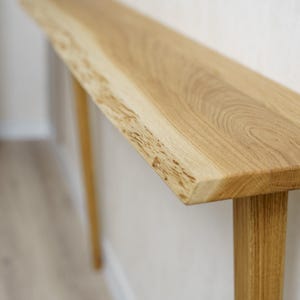 May include: A light-colored wooden console table with a natural edge on the top. The table has a rectangular top and two legs. The wood grain is visible. The table is approximately 60 inches long.