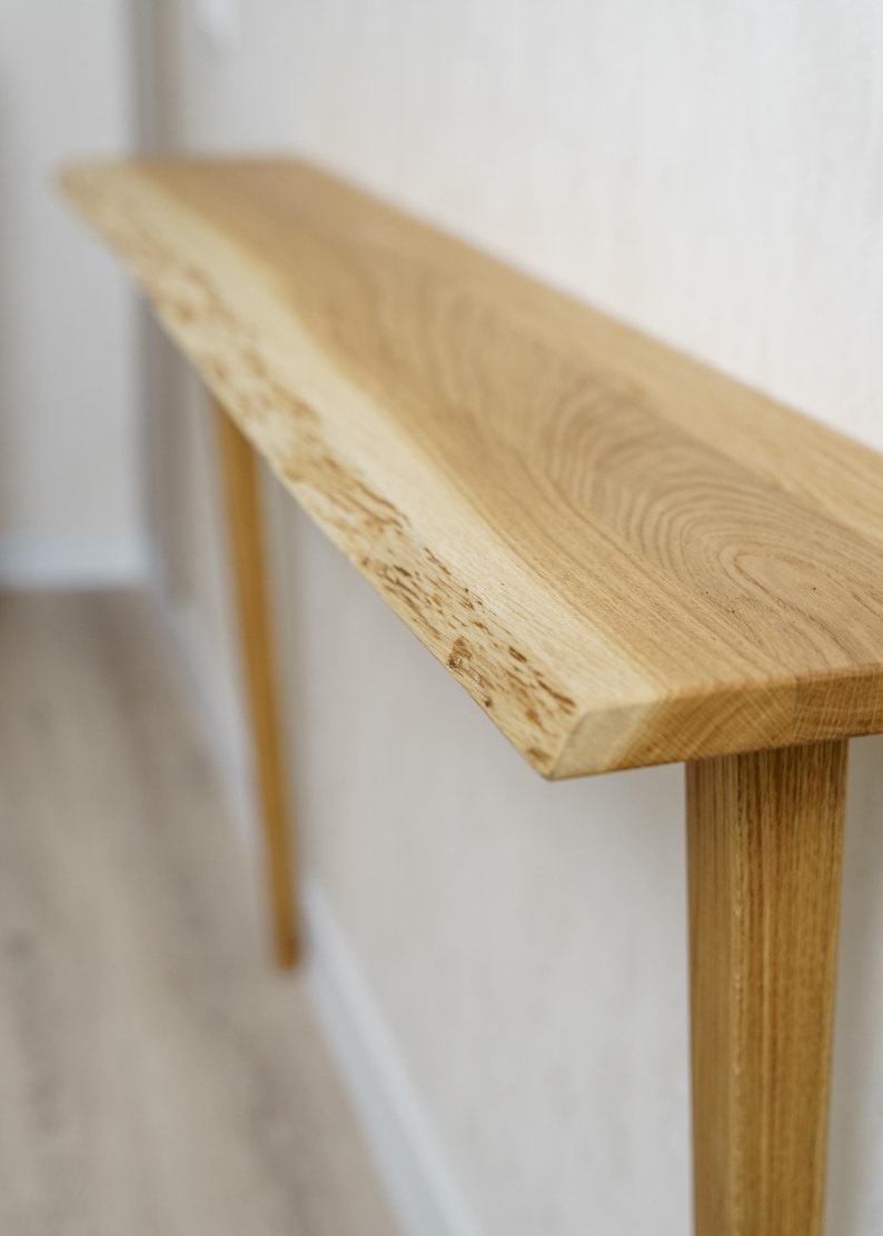 May include: A light-colored wooden console table with a natural edge on the top. The table has a rectangular top and two legs. The wood grain is visible. The table is approximately 60 inches long.