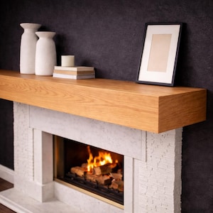 May include: A contemporary fireplace featuring a light oak mantel and a white stone surround. Two white vases, a candle, and a framed picture are on the mantel. The fireplace has a burning fire.