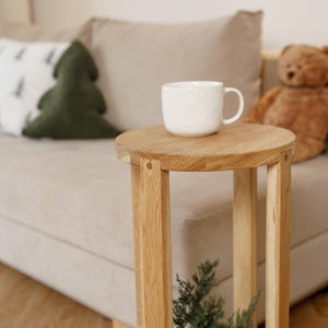 Puede incluir: Una taza de cerámica blanca con un diseño floral se encuentra sobre una pequeña mesa auxiliar redonda de madera. La mesa tiene un acabado de madera natural y está sostenida por cuatro patas verticales. Una almohada verde en forma de árbol de Navidad y un oso de peluche están en el fondo.