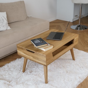 Könnte beinhalten: Ein hellbrauner Couchtisch aus Holz mit einer Schublade und einem unteren Regal. Ein Laptop und zwei Bücher liegen auf dem Tisch, der auf einem weißen, flauschigen Teppich steht. Der Tisch hat vier angewinkelte Beine.