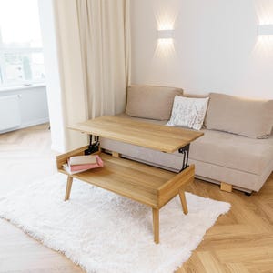 May include: A light-colored wooden lift-top coffee table with a lower shelf, set on a white shag rug. The table is in a living room setting with a beige sofa and a window with white curtains. Two books are on the lower shelf.