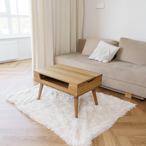 Könnte beinhalten: Ein Couchtisch aus hellem Holz mit rechteckiger Platte und vier abgewinkelten Beinen. Der Tisch hat eine eingelassene Fläche auf der Oberseite. Er steht auf einem weißen, flauschigen Teppich. Ein beiges Sofa und Vorhänge sind im Hintergrund.