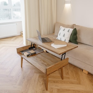 Puede incluir: Una mesa de centro de madera clara con tapa elevable, un portátil, libros y un estante inferior para almacenamiento. La mesa está en un salón con un sofá beige, cojines y una ventana con cortinas.
