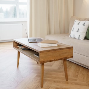 May include: A light oak coffee table with a rectangular top and angled legs. A laptop and books rest on the table's surface. The table has a lower shelf for storage. The room has a neutral colour palette.