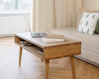 Puede incluir: Una mesa de centro de roble claro con una parte superior rectangular y patas anguladas. Una computadora portátil y libros descansan sobre la superficie de la mesa. La mesa tiene un estante inferior para almacenamiento.