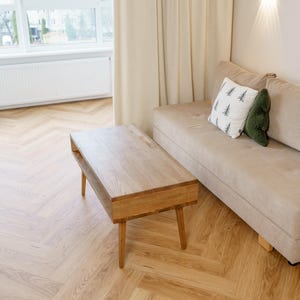 May include: A living room scene featuring a light-colored sofa with decorative pillows and a wooden coffee table. The table has a rectangular top and tapered legs. The room has light-colored curtains and a herringbone wood floor.