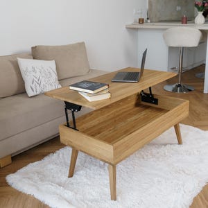 May include: A light-colored wooden lift-top coffee table with a lower storage compartment. The table is on a white fluffy rug. A laptop and books are on the table. A beige sofa and a white wall are in the background.