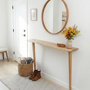 Puede incluir: Una consola de madera clara con una encimera de borde vivo, un espejo redondo y una cesta de mimbre llena de toallas. Un jarrón con flores está sobre la mesa. Una alfombra gris y botas marrones están en el suelo.