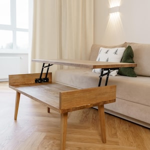 May include: A light brown wooden lift-top coffee table with a bench-style base. The table has a rectangular top that can be raised. The legs are tapered. The table is in a living room setting with a beige sofa and light-colored curtains.